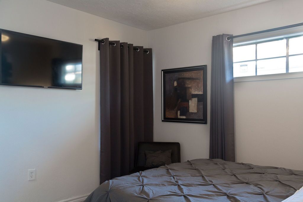 A bedroom with a bed, a flat-screen TV, a painting, and a window with curtains.