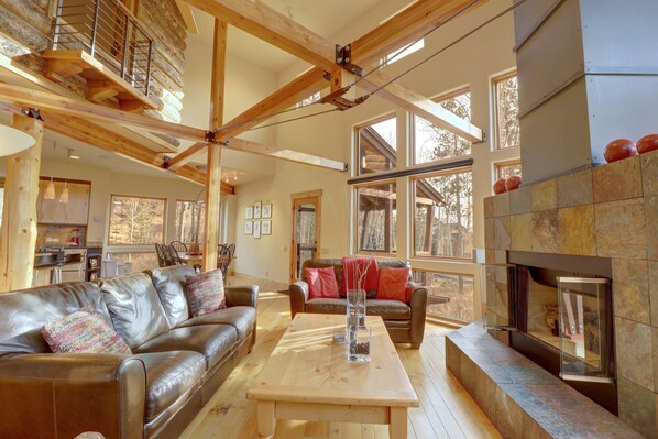 A spacious living room with a fireplace, wooden beams, and large windows.