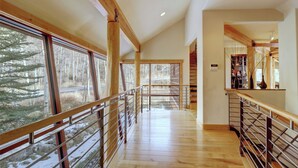A spacious hallway with wooden floors and metal railings, large windows offering a view of trees, and a staircase leading to another level.