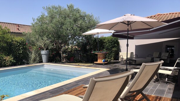 Une piscine entourée de verdure, avec des chaises longues et un parasol, et un patio carrelé.