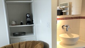 Une salle de bain avec un lavabo blanc, un miroir et un porte-serviettes. On y trouve une étagère avec une machine à café et une tasse.