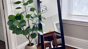 Una planta en maceta junto a un espejo con un atril, una silla de madera y una ventana con persianas.