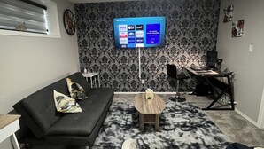 Ein Wohnzimmer mit schwarzem Sofa, Couchtisch, Wandfernseher, einem kleinen Beistelltisch und einem gemusterten Teppich.