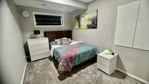 Ein Schlafzimmer mit Bett, Nachttisch, Kommode, Wanduhr und Fenster.