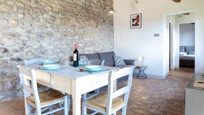 A dining area with a stone wall, a table set with plates, glasses, and a bottle of wine. There is a sofa with cushions and a small side table with a lamp.