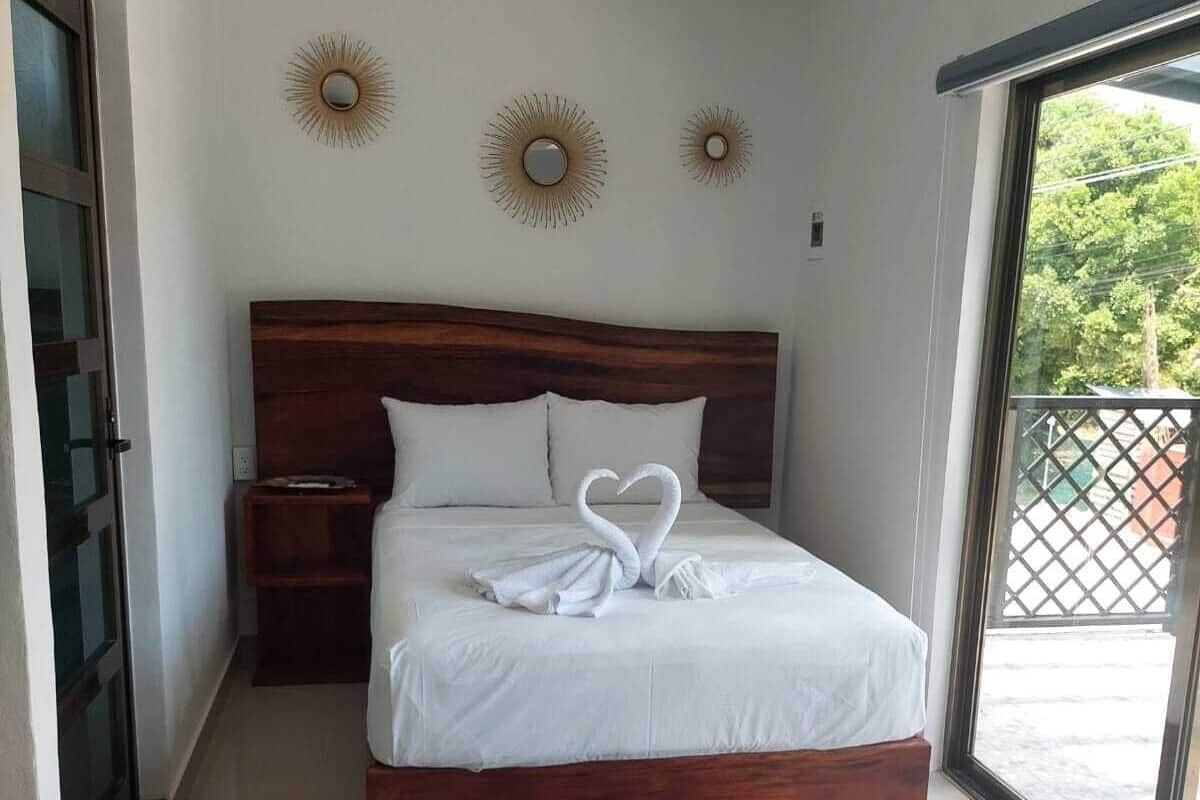 A bedroom with a wooden bed, white bedding, and a heart-shaped towel on the bed. There are two blue rugs on the floor, a ceiling fan, and a window with a view of greenery.