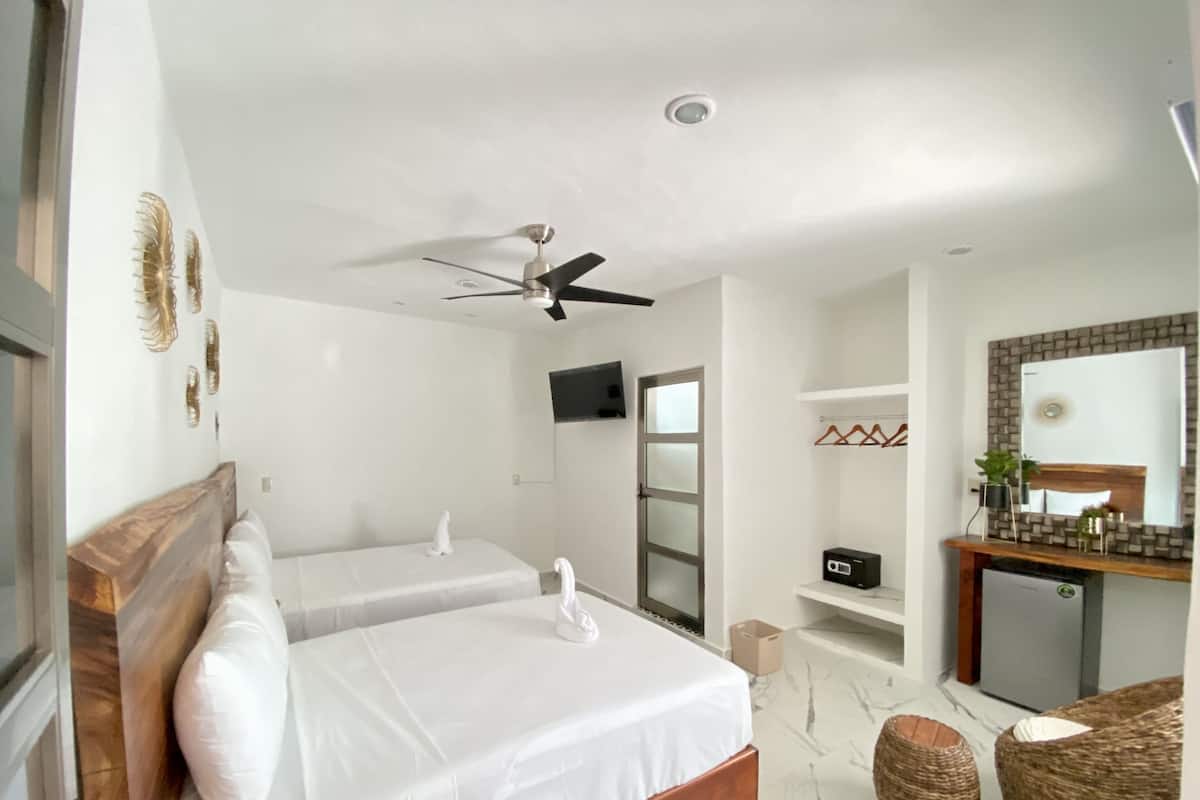 A hotel room with two beds, a ceiling fan, a TV, a mirror, a small kitchenette, and a bathroom with a shower.