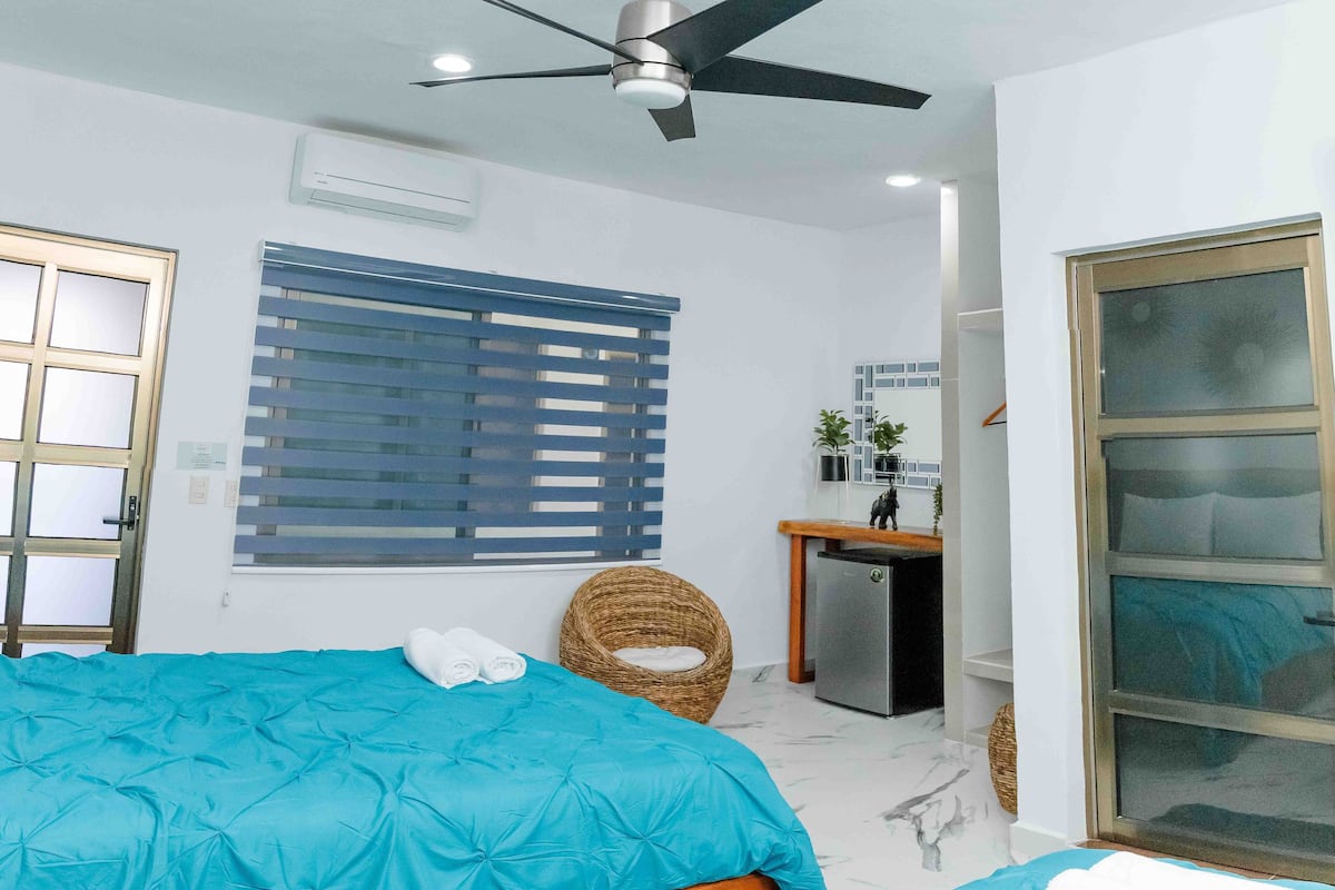 A bedroom with a bed, a ceiling fan, a window with blinds, a small desk with a refrigerator, and a door with a glass panel.