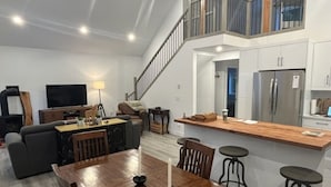 A modern living room with a wooden dining table, a sofa, and a TV. There is a kitchen area with a refrigerator and a wooden countertop.