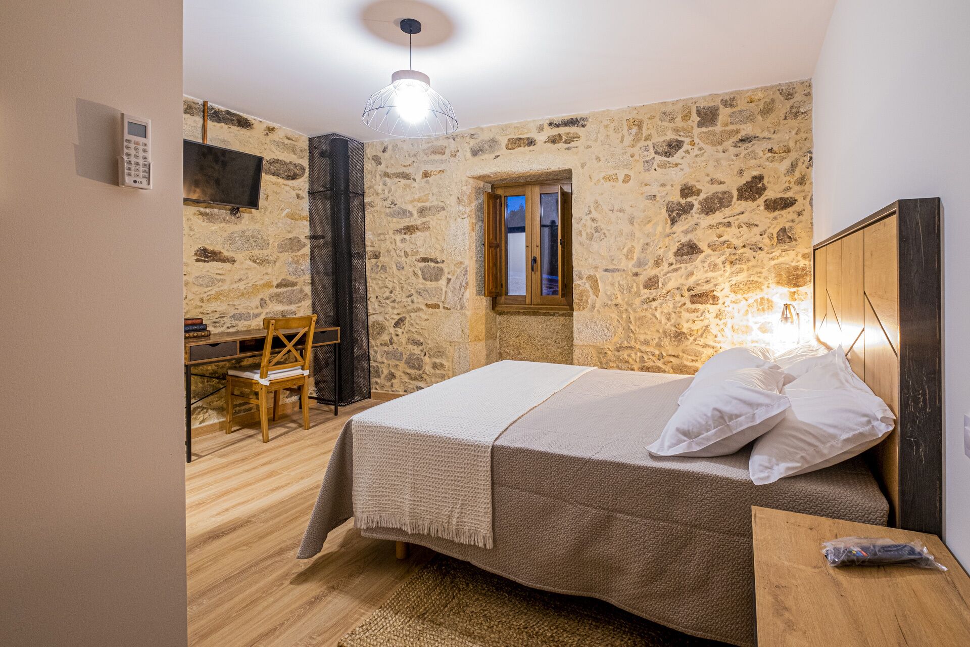 A bedroom with a stone wall, a bed with white bedding, a wooden desk, and a chair.