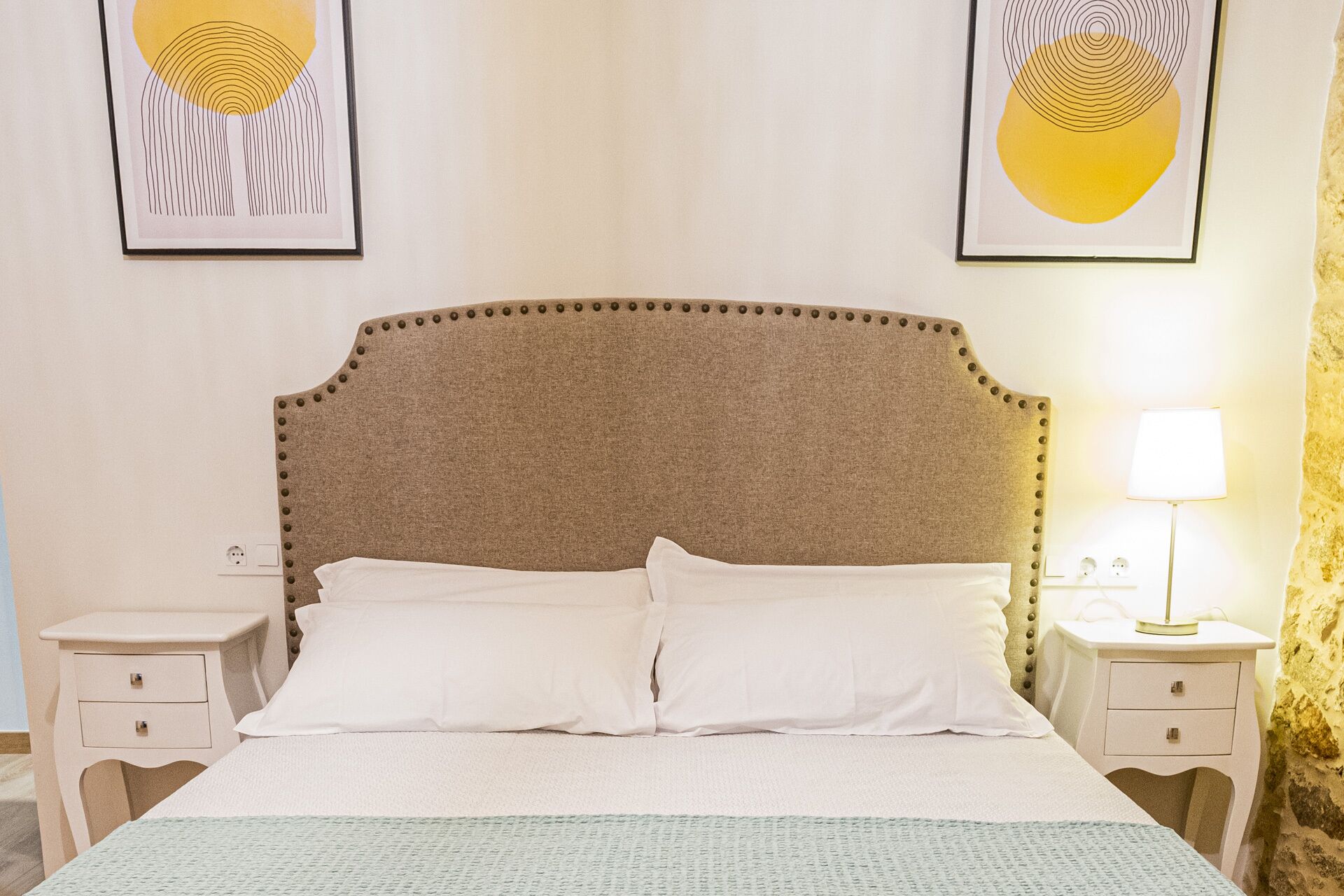 A neatly made bed with a textured headboard, two bedside tables with lamps, and two framed abstract paintings on the wall.