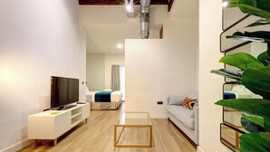 A modern living room with a flat-screen TV, a coffee table, and a sofa. There is a bed in the background and a staircase with a plant on the right. - Heredia Suites Centro (Málaga)
