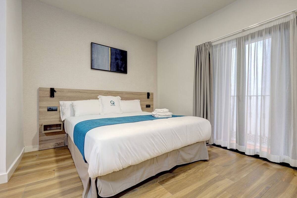 Une chambre d’hôtel bien aménagée, avec une tête de lit en bois, un lit recouvert de draps blancs agrémentés d’une touche de bleu, une petite table de chevet avec une lampe et une fenêtre ornée de rideaux légers.
