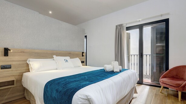 A modern hotel room with a large bed, a wooden headboard, a balcony with a view, and a red chair. - Heredia Suites Centro (Málaga)