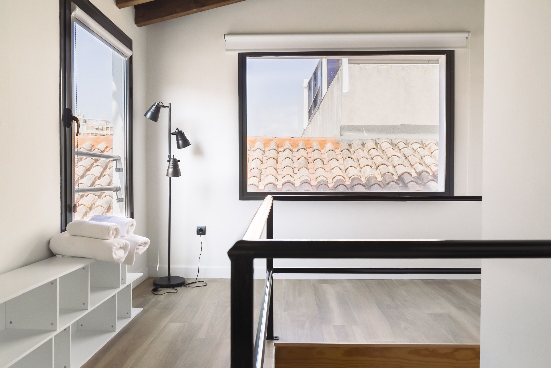 Una habitación moderna con un ventanal grande, un banco blanco con toallas, una lámpara de pie y una estructura de cama de metal negro.