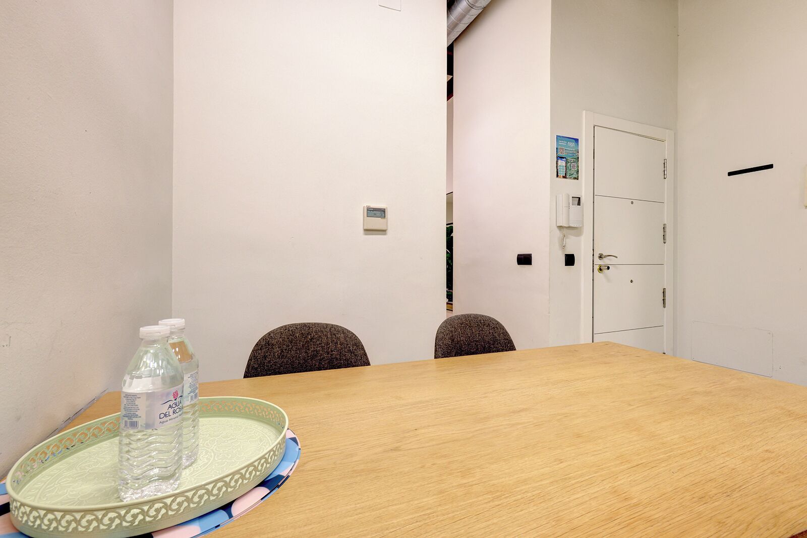 A conference room with a wooden table, two chairs, a water bottle on a tray, and a door with a phone and a poster on the wall.