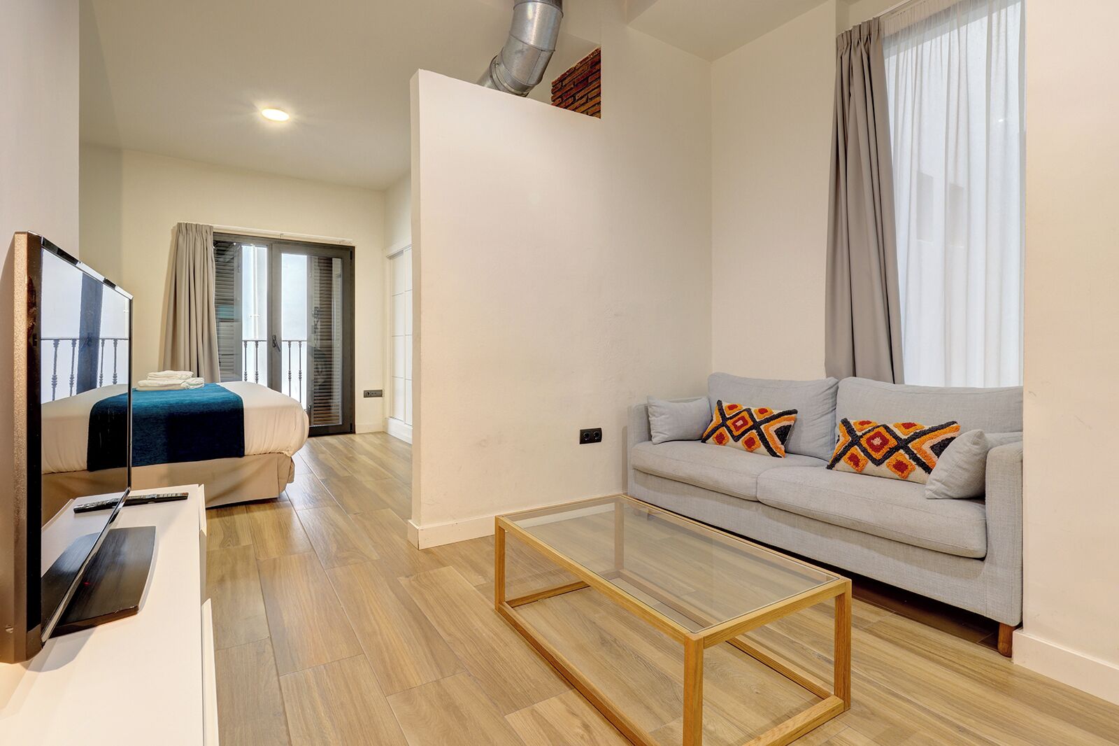 A modern hotel room with a glass coffee table, a sofa with patterned cushions, a bed with a blue blanket, and a TV on a stand.