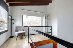 Un intérieur moderne avec un plancher en bois, une chaise grise, une rambarde en métal noir et une fenêtre donnant sur un toit recouvert de tuiles.