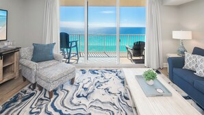 Una sala de estar con un gran ventanal corredizo a un balcón con vista al océano, un sofá azul, un sillón estampado y una mesa de centro con una planta.