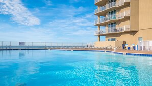 Una piscina con un cielo azul despejado, un edificio de varios pisos con balcones y una valla a lo largo del borde de la piscina.