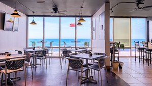 Un comedor interior con mesas y sillas, ventanas amplias con vista a la playa y ventiladores de techo.