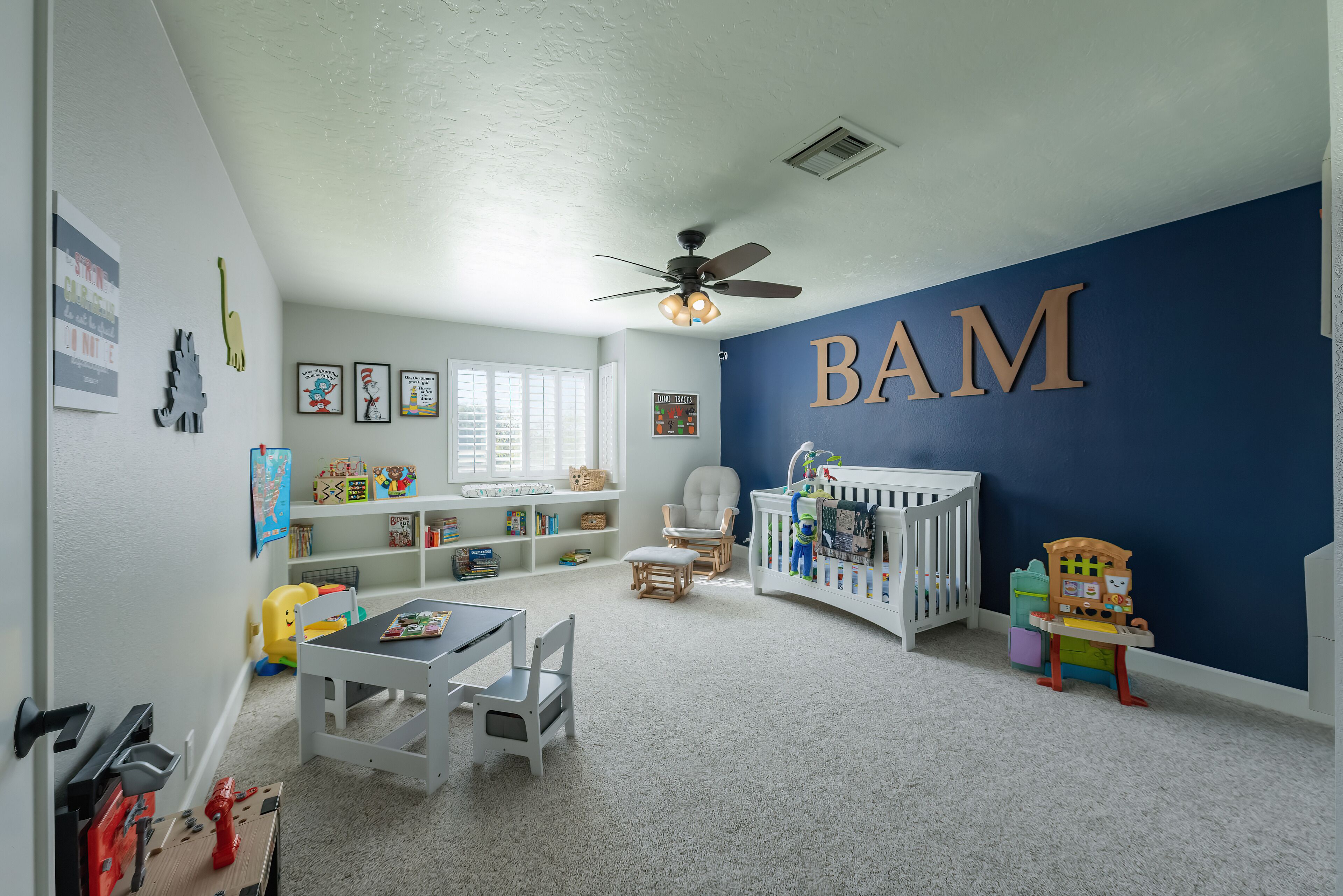 A well-lit room with a ceiling fan, a crib, a bookshelf, and a play area with toys and a table.