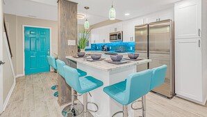 Une cuisine moderne avec des meubles blancs, un îlot central doté de tabourets de bar, des appareils en acier inoxydable et une porte de couleur turquoise.