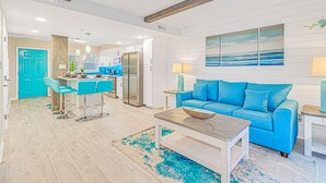 Un salon moderne avec un canapé bleu, une table basse et un coin cuisine doté de meubles blancs et d’une porte bleue.