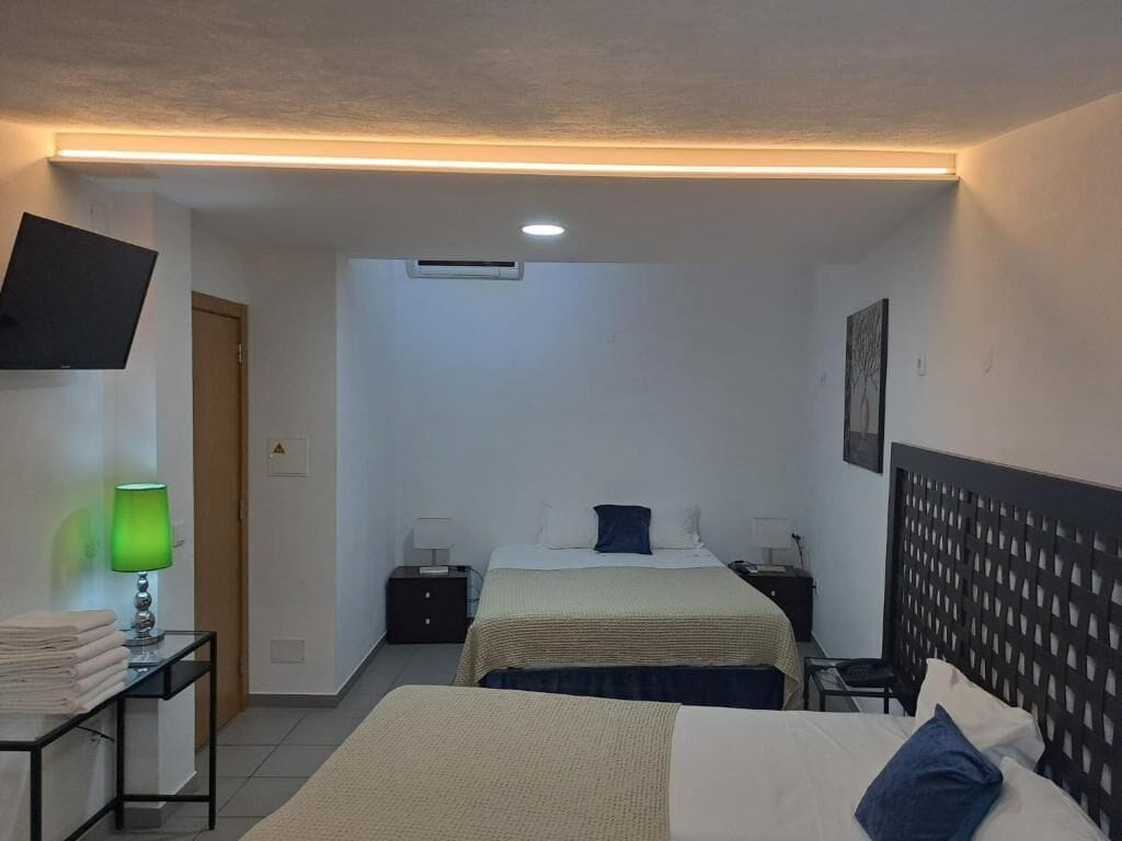 Camera d'albergo con due letti, una TV a schermo piatto montata a parete, un comodino con una lampada e un tavolino con asciugamani piegati.