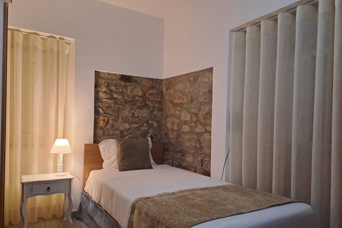 Ein Schlafzimmer mit einer Wand aus Stein, einem Bett mit weißer und brauner Bettdecke, einem Nachttisch mit Lampe und einem Fenster mit vertikalen Jalousien.