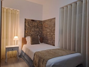 Un dormitorio con pared de acento de piedra, una cama con cubrecama blanco y marrón, una mesita de noche con lámpara, y una ventana con persianas verticales.