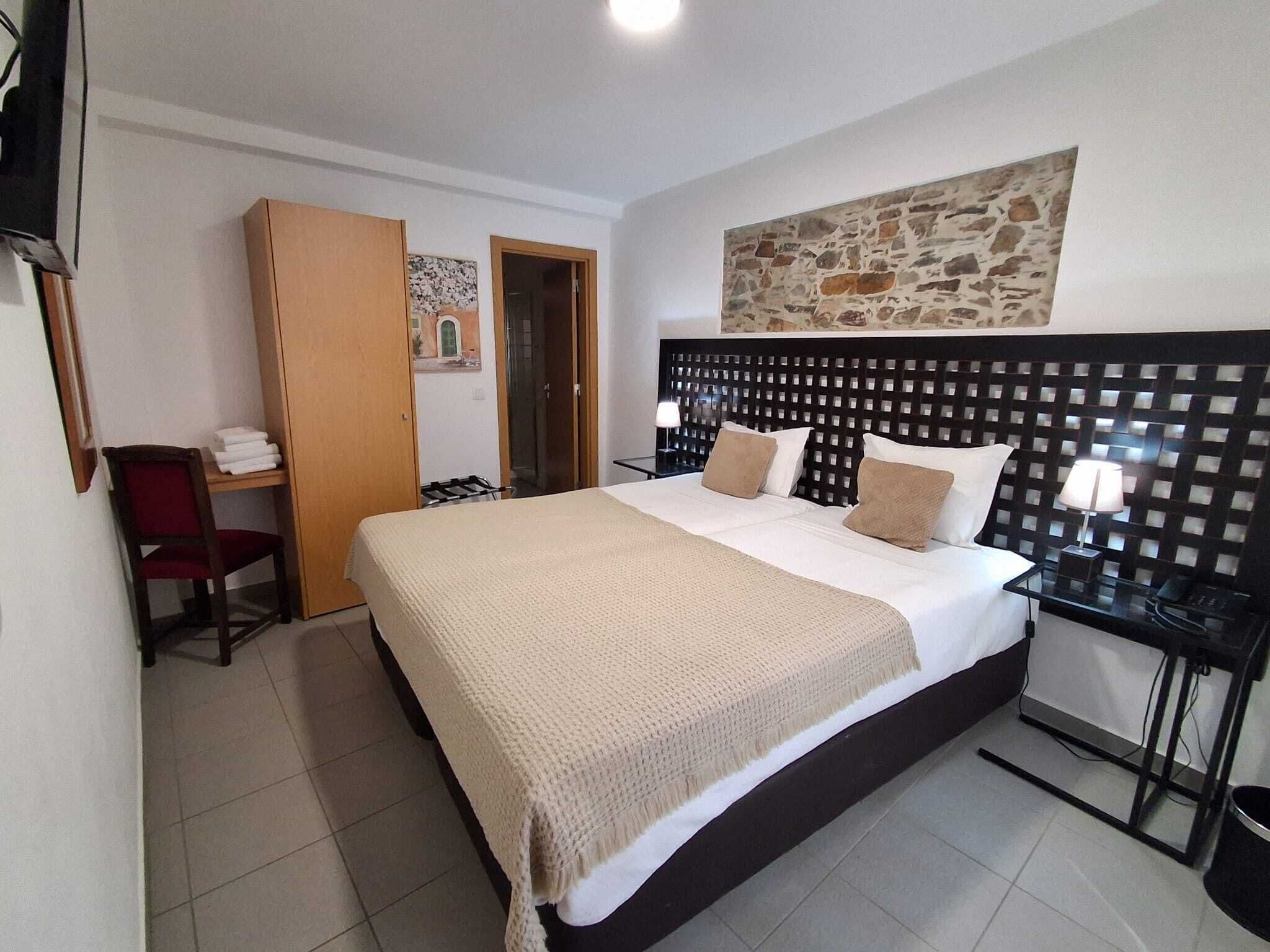 Quarto de hotel com uma cama grande, uma escrivaninha, uma cadeira, uma mesinha de cabeceira e uma parede de pedra.