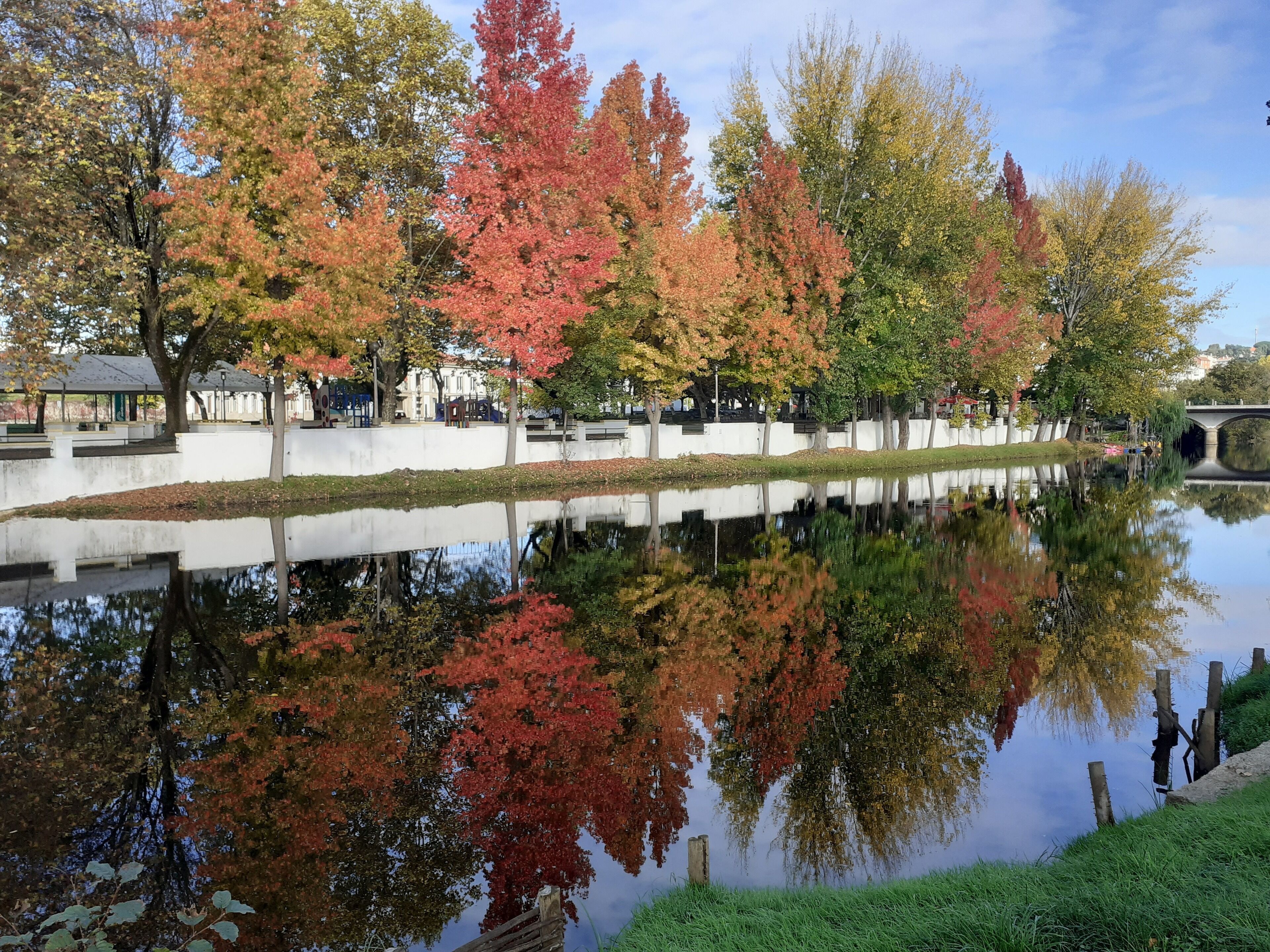 A calm water body reflecting trees with autumn foliage, a white wall, and a bridge in the background.