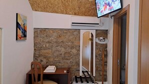 Una habitación con pared de piedra, puerta de madera, televisor montado en la pared, una silla, una mesa con una toalla y una estantería metálica.