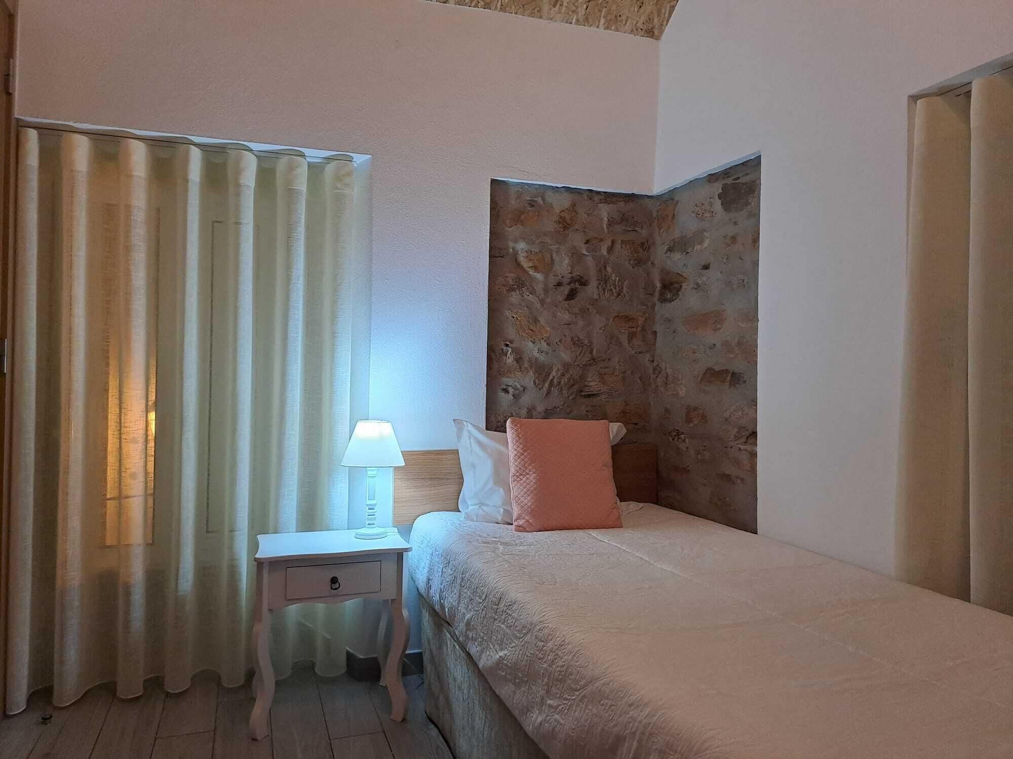 A bedroom with a bed, a nightstand with a lamp, a window with curtains, and a stone wall.