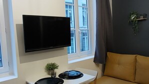 A modern living room with a sofa, a TV mounted on the wall, a dining table with chairs, and a window with curtains. - Renovated studio 5 minutes from the city center (Rouen)
