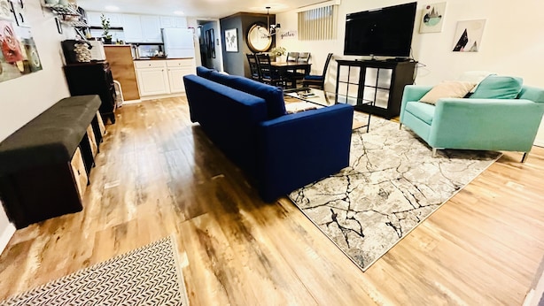 A living room with a blue sofa, a teal armchair, and a coffee table. There is a kitchen in the background with white cabinets and a dining area with a table and chairs.