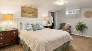 A bedroom with a large bed, two bedside tables with lamps, a chair, a wardrobe, a window with blinds, and a plant.