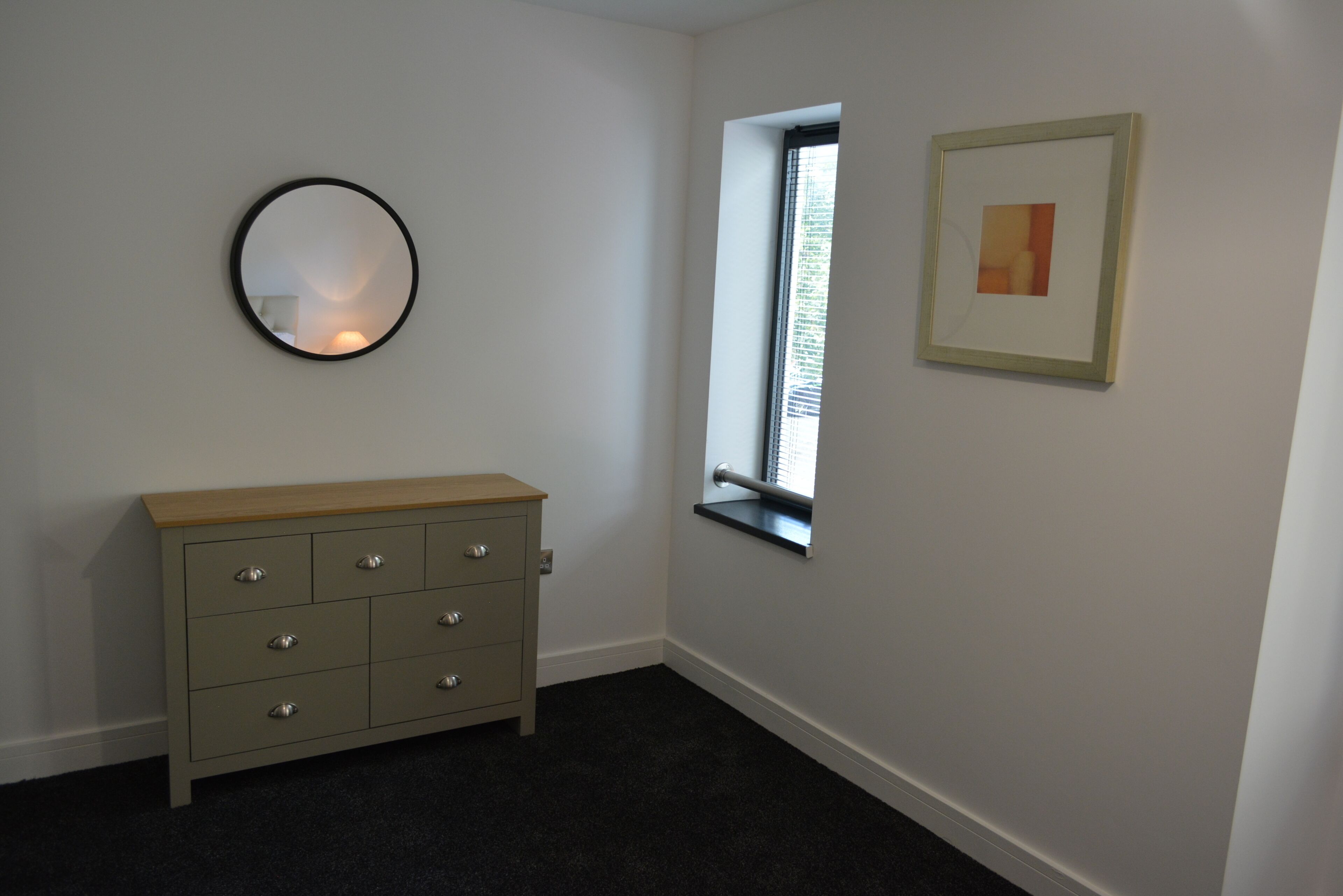 Quarto com penteadeira, espelho redondo, janela e quadro na parede.