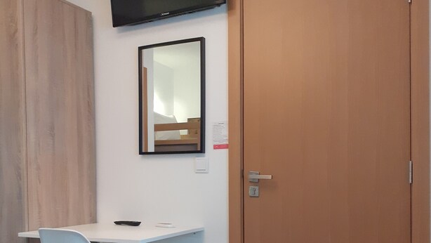 A room with a wooden wardrobe, a desk with a chair, a flat-screen TV mounted on the wall, and a wooden door. - Hostel Sertã (Serta)