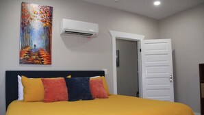 Quarto de hotel com uma cama, almofadas coloridas, um quadro na parede, um aparelho de ar condicionado e uma porta branca.