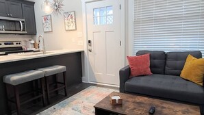 Cozinha moderna com armários escuros, micro-ondas e pia. Há um espaço com banquetas, um sofá cinza com almofadas vermelhas e amarelas, e uma mesa de centro com um controle remoto.