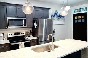 A modern kitchen with dark cabinets, stainless steel appliances, a white countertop, and pendant lighting. - West End Jewel on Sunset (Winston-Salem)