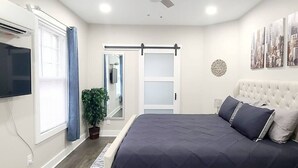 A bedroom with a bed, a TV, a sliding door, a plant, and artwork on the wall. - West End Jewel on Sunset (Winston-Salem)