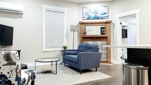 A modern living room with a sofa, a coffee table, and a TV. There is a fireplace with a decorative mantel and a large window with blinds. - West End Jewel on Sunset (Winston-Salem)