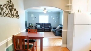 A modern kitchen with a wooden dining table and chairs, a white refrigerator, and a living area with a sofa and a ceiling fan. - Cozy Ardmore Cottage Main Level (Winston-Salem)