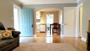 A living room with a brown leather sofa, a beige carpet, and a blue door leading to a kitchen area with wooden cabinets and a dining table. - Cozy Ardmore Cottage Main Level (Winston-Salem)