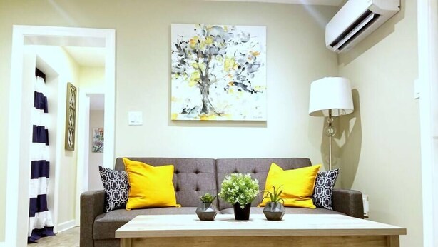 Un salon avec un canapé gris, des coussins jaunes, une table basse en bois et un tableau représentant un arbre.