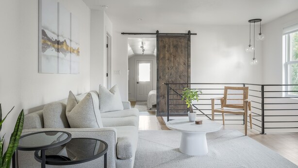 A modern living room with a grey sofa, a coffee table, and a plant. There is a sliding barn door and a view of a balcony with a railing. - Proper Luxury, Sauna, The Woodland Loft (Bozeman)