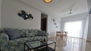 A living room with a sofa, a glass coffee table, a dining area with a table and chairs, and a ceiling fan.